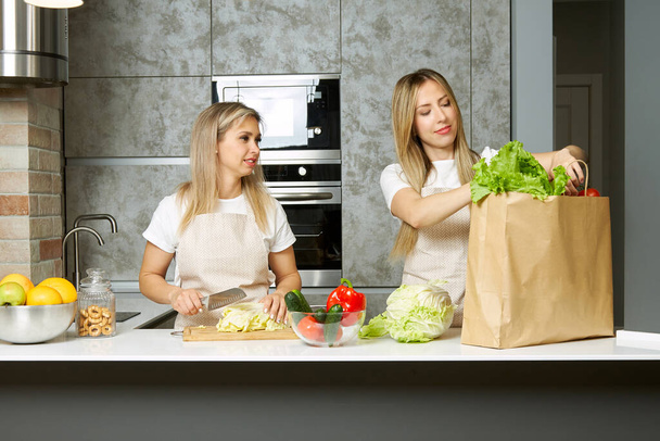 Two young smiling women take apart a paper bag of healthy foods and prepare vegetarian vegan food in the modern kitchen of the house - Valokuva, kuva