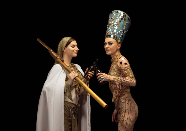 Young beautiful woman in image of Nefertiti in art performance with her servant drinking beer isolated on dark background. Retro style, comparison of eras, humor concept. - Foto, Imagem
