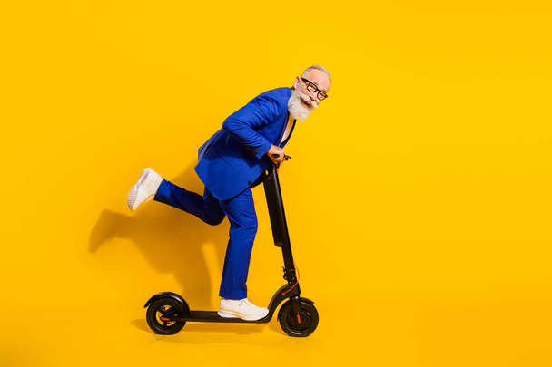 Full size profile side photo of happy smiling good mood mature man riding eco scooter isolated on yellow color background - 写真・画像