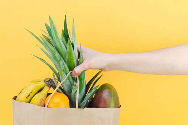 Paper shopping bag filled with tropical fruit with woman's hand on yellow background. Copy space. - Valokuva, kuva