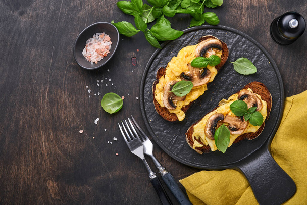 Scrambled eggs with fried mushrooms and basil on bread on black table background. Homemade breakfast or brunch meal - scrambled eggs and mushrooms sandwiches. Top view with copy space - Photo, image