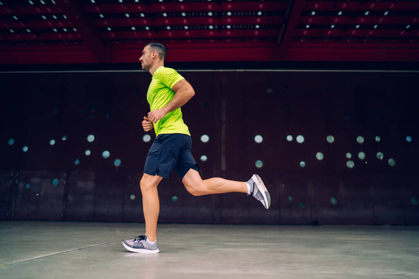 Caucasian active man dressed in sportswear jogging during morning workout losing weight at urban setting, side view of male athlete 30s training endurance and physical strength on cardio running - Fotografie, Obrázek