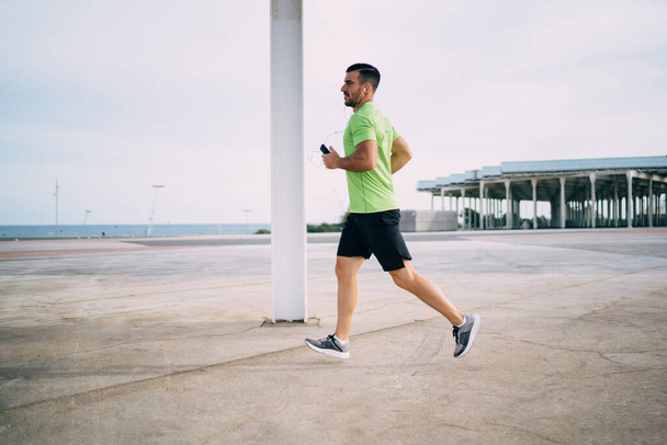 Side view of determined male athlete in electronic headphones listening music songs during cardio running around city, Caucaisan sportsman in modern earphones running during physical recreation - Foto, imagen