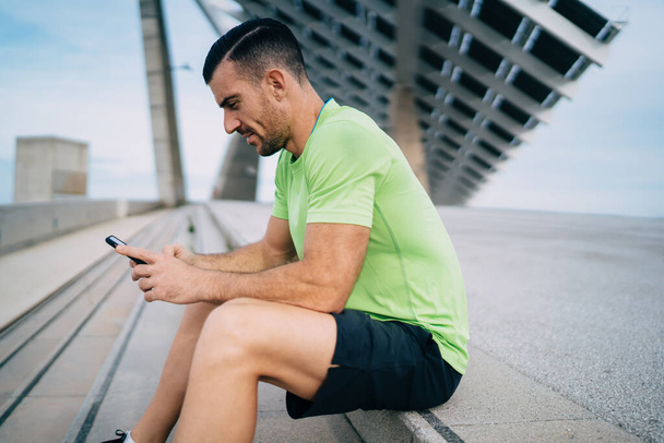 Side view of young Caucasian sportsman reading motivation publication during cellphone networking , muscular male athlete checking received email on cellular technology with 4g wireless connection - Fotografie, Obrázek