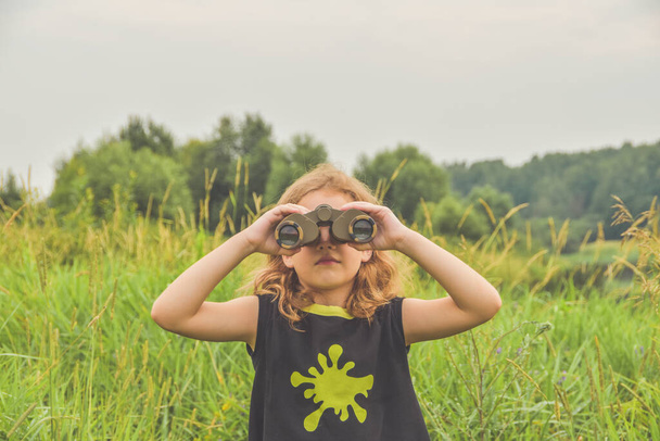 the child girl stands in a meadow and looks through binoculars - Foto, Imagen