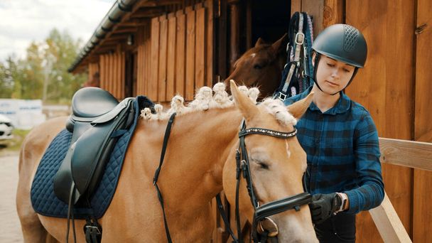 Jockeyspielerin fixiert die Waage ihres hellbraunen Pferdes außerhalb des Stalls - Foto, Bild