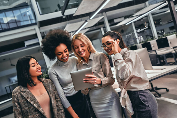 Group of beautiful young business women standing in business center holding tablet in hands and laughing. - Fotoğraf, Görsel