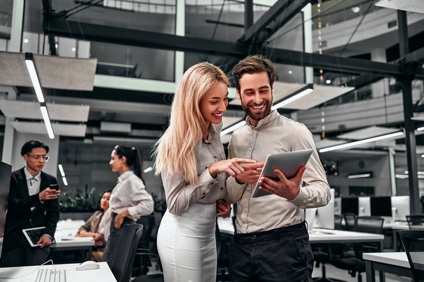 Two beautiful successful smiling business people are standing looking at the tablet screen in a spacious office. Colleagues are working in the background. - Foto, imagen