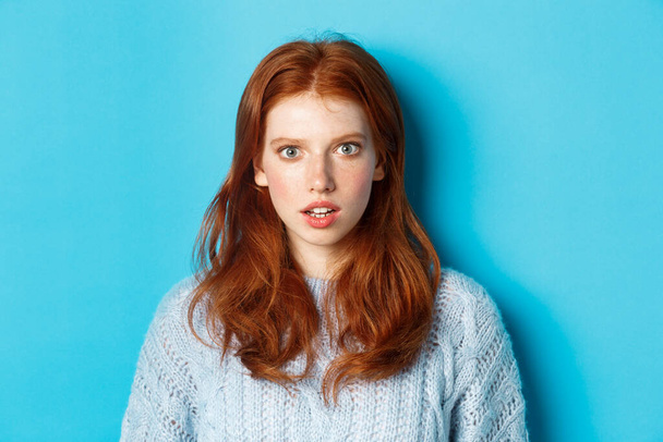 Close-up of amazed redhead girl staring at camera with complete disbelief, standing shocked against blue background - Foto, afbeelding