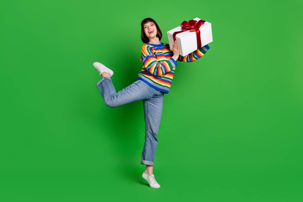 Full length photo of funny carefree young woman wear striped sweater smiling giving gift box isolated green color background - Foto, Bild