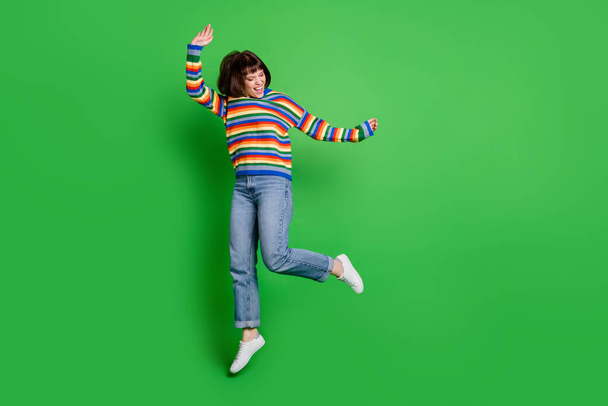 Full length photo of pretty funky young woman wear striped sweater smiling jumping high isolated green color background - Foto, imagen