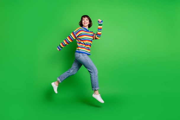 Full length photo of funky sweet young woman wear striped sweater smiling jumping high running isolated green color background - Foto, afbeelding