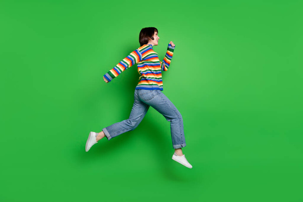 Full length photo of sweet pretty young woman wear striped sweater smiling jumping running empty space isolated green color background - Fotoğraf, Görsel