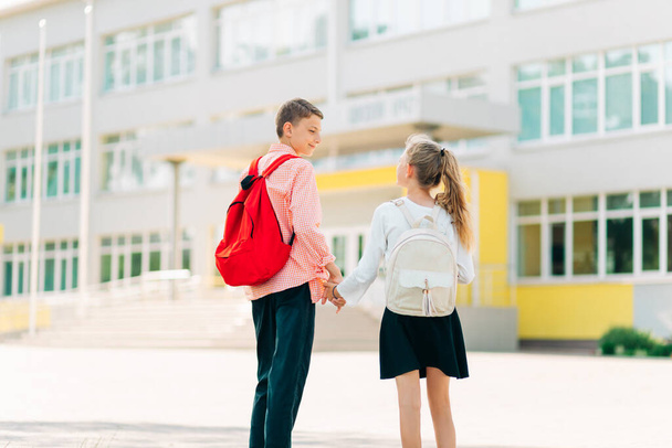Back to school, Two happy children are ready for primary school, pupils on the first day of school, a boy and a girl go hand in hand to lessons with backpacks, near the school building - Φωτογραφία, εικόνα