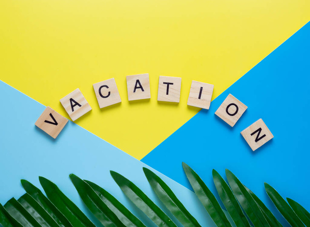 small wooden cubes with letters on a yellow-blue background with leaves from plma. The word VACATION. concept of rest at sea. copy space. - Foto, imagen