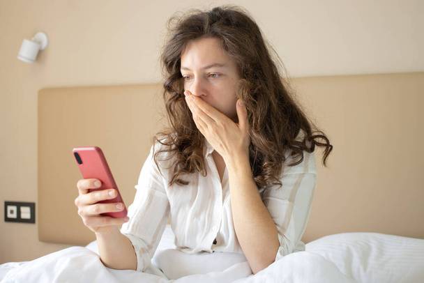 Jeune femme choquée regardant smartphone, assis dans un lit confortable blanc à la maison, lisant des nouvelles inquiétantes dans les médias, regardant du contenu vidéo, se sentant frustré par des informations inattendues. - Photo, image