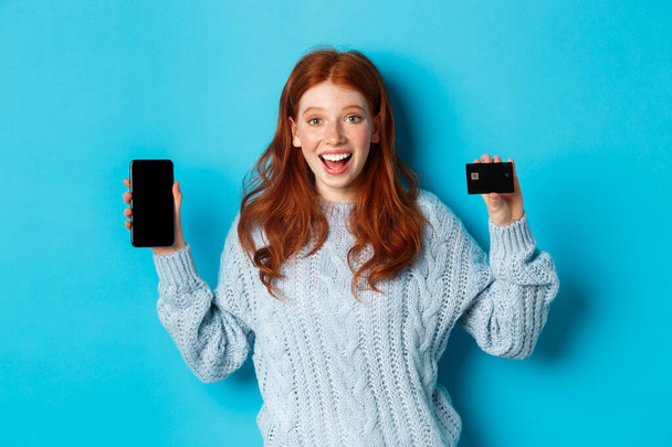 Heyecanlı kızıl kız cep telefonu ekranı ve kredi kartı gösteriyor, online mağaza veya uygulama gösteriyor, mavi arka planda duruyor. - Fotoğraf, Görsel