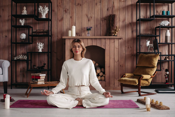 Jovencita sonriente practicando yoga sentada en pose de loto, meditando en el acogedor interior del hogar. Entrenamiento femenino para el bienestar - Foto, imagen