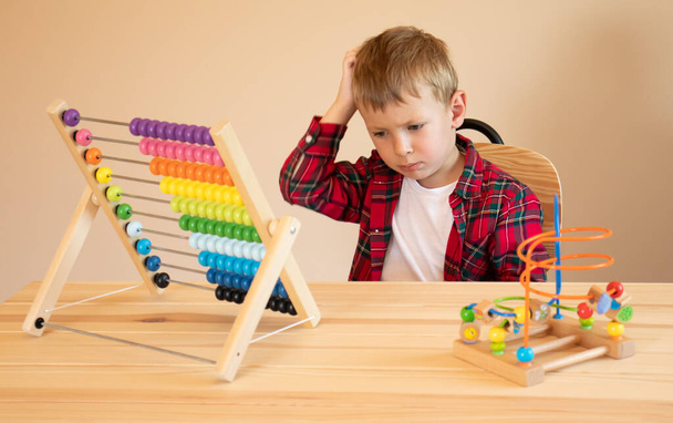 Маленький хлопчик сидить за столом і грає з дерев'яними освітніми іграшками, подряпає голову і думає про завдання
. - Фото, зображення