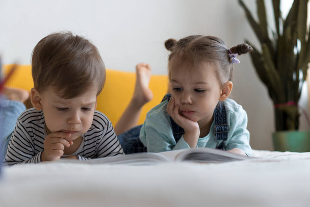 Two preshool, toddler children read large interesting book of fairy tales on yellow bed. Siblings little twins reader have fun, happy kid on quarantine at home. Friendship, family, education concept - Foto, Imagem