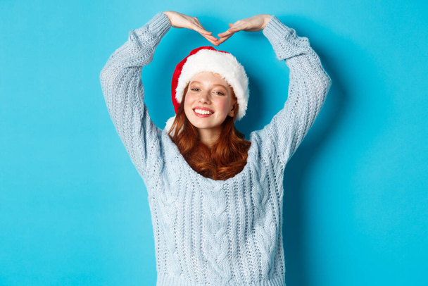 Kış tatili ve Noel arifesi konsepti. Noel Baba şapkası ve süveteri içinde sevimli kızıl saçlı genç kız, kalp işareti yapıyor ve gülümsüyor, neşeli xmas diliyor, mavi arka planda duruyor. - Fotoğraf, Görsel