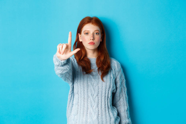 Σοβαρή κοκκινομάλλα με πουλόβερ που δείχνει χειρονομία ταμπού, απλώνοντας το ένα δάχτυλο, κουνώντας το δείκτη για να αποδοκιμάζει, διαφωνεί και απαγορεύει κάτι, μπλε φόντο - Φωτογραφία, εικόνα