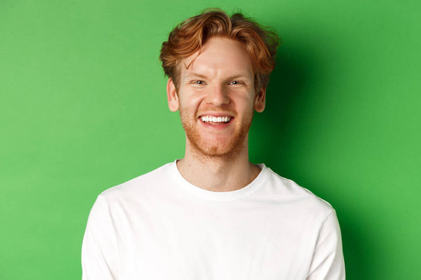 Emotionen und Modekonzept. Glücklicher junger Mann mit roten Haaren und Bart, lächelnd und lachend in die Kamera, vor grünem Hintergrund stehend - Foto, Bild