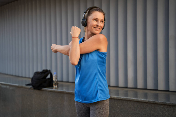 Αθλητική ώριμη γυναίκα σε αθλητικά και ακουστικά χαμογελώντας ενώ ζεσταίνεται, τεντώνει τα χέρια της, στέκεται σε εξωτερικούς χώρους - Φωτογραφία, εικόνα