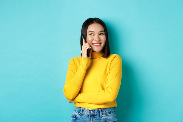 Attractive korean woman in yellow sweater, having conversation and smiling, talking on mobile phone, looking left at copy space, standing over blue background - Photo, Image
