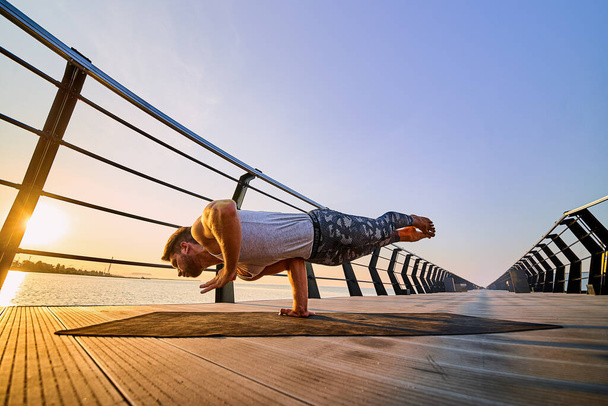 Fit man doing a one hand stand while practicing yoga alone near the ocean against sky at dusk or dawn - Foto, immagini