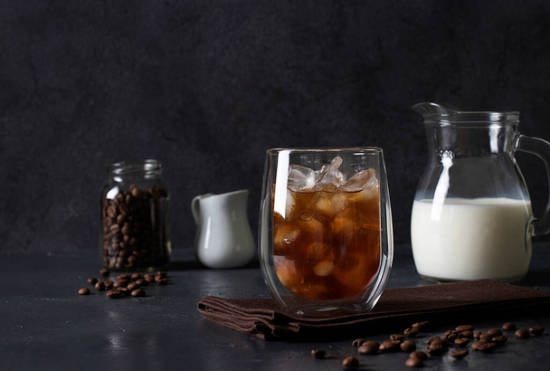 Kalter Kaffee in einem Glas mit Eiswürfeln, in das anschließend Milch gegossen wird, dunkelgrauer Hintergrund - Foto, Bild