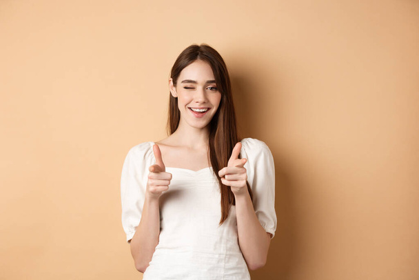 We need you. Smiling young woman pointing fingers at camera to beckon or invite people, standing on beige background - Photo, Image