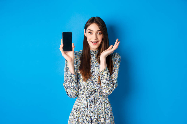Izgalmas lány bemutató üres okostelefon képernyő és lélegzetelállító lenyűgözte, demonstrálja bevásárló alkalmazás, álló kék háttér - Fotó, kép