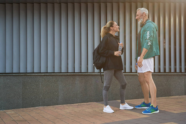 Zijaanzicht van sportieve mensen van middelbare leeftijd in sportkleding die naar elkaar kijken, praten terwijl ze samen buiten staan na fitnesstraining - Foto, afbeelding
