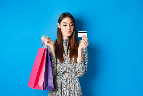 美しい女の子はプラスチッククレジットカードで買い物に行く,購入したアイテムと紙の袋を保持,青の背景に満足立って - 写真・画像