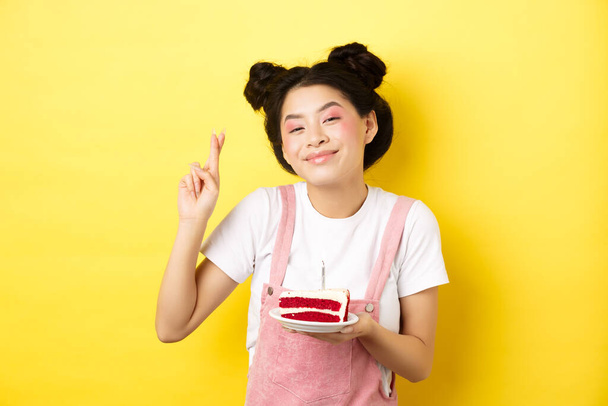 Ünnepek és ünneplés. Pozitív ázsiai szülinapos lány keresztujjú, kívánság b-napos torta és gyertyagyújtás, mosolygós boldog a kamera, sárga háttér - Fotó, kép