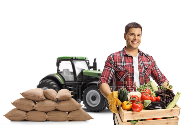 Young farmer with a tractor and pile of sacks holding a crate with vegetables isolated on white background - Fotó, kép