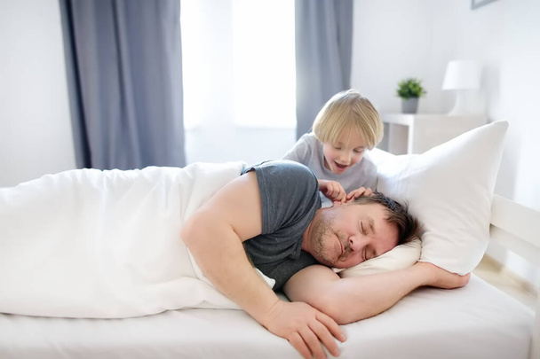 Little child awake. Early in the morning he wakes up his sleeping father in bed at home. Tired dad hears nothing, sleeps and does not wake up. Parenthood. Tired and burnout parents. - Foto, Imagen