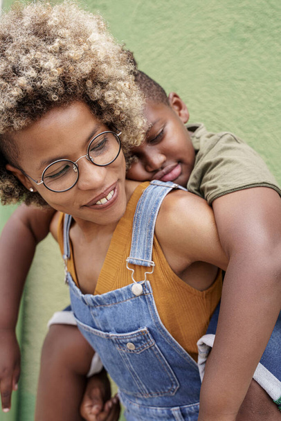 Black Loving Mother piggybacking sleepy cute little black Son in front of green wall - Photo, Image