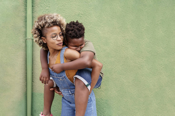 African American Young Loving Mother piggybacking sleepy cute little black Son in front of green wall - Fotografie, Obrázek