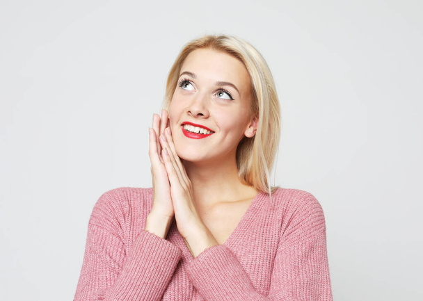 Smiling blond woman wearing pink sweater touching perfect skin and smiling, standing on grey background. - 写真・画像