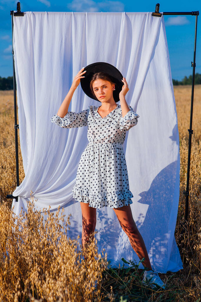 Young beautiful woman dressed in a white Polka-dot dress and black hat standing at golden oat field with a white curtain on the background. - Fotoğraf, Görsel