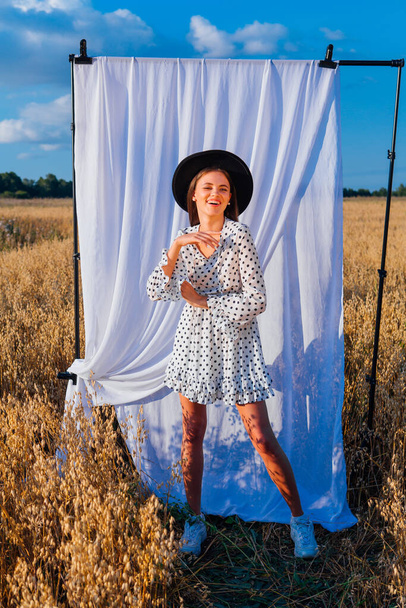 Young beautiful woman dressed in a white Polka-dot dress and black hat standing at golden oat field with a white curtain on the background. - Zdjęcie, obraz