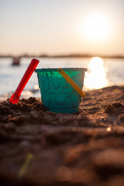 Kinderspeelgoed om op het zand te spelen. Plastic emmer en hark op het strand bij zonsondergang. Het concept van zomer, gezinsvakantie en vakantie. - Foto, afbeelding