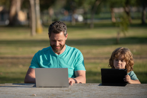 On line learning. Father and son with laptop studying online at green summer park. Little kid sitting at the table and watching online lesson on notebook, daddy helping child to do homework. - Zdjęcie, obraz