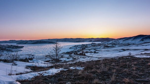 Talven auringonnousu Siperiassa. Vuorten yläpuolella oleva taivas on vaaleanpunainen ja oranssi. Auringon hehku jäätyneen järven jäällä. Kylän talot rannalla. Lunta ja kuivaa ruohoa maassa. Baikal - Valokuva, kuva