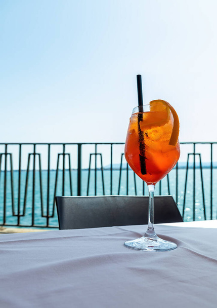 Sklenice ledově studeného koktejlu Aperol spritz podávaná ve sklenici na víno, zdobená plátky pomeranče, umístěná na stole s výhledem na moře. Letní středomořská dovolená.  - Fotografie, Obrázek