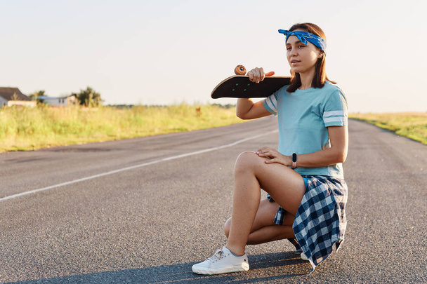 Young adult attractive girl with dark hair posing with a longboard over shoulders while sitting on the road in summer, looking away with pensive expression. - Foto, immagini