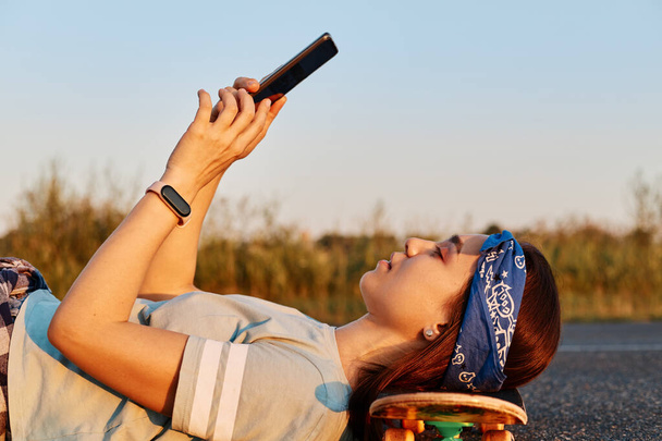 Side view portrait of slim beautiful woman wearing hair band and t shirt laying on asphalt road and keeping head on skateboard, holding phone in hands, making selfie or browsing internet. - Photo, image