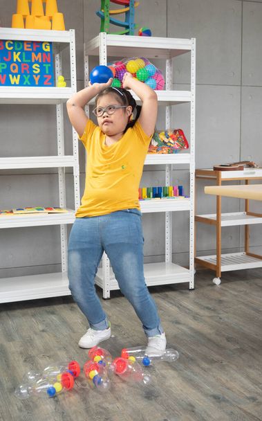 Una linda niña con síndrome de Down apuntando antes de lanzar una pelota de plástico al blanco mientras juega juegos en el aula especial para niños. - Foto, imagen